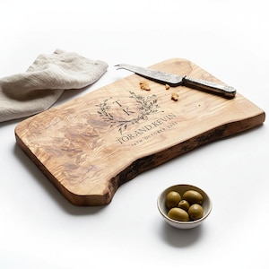 May include: A wooden serving board with a natural edge, engraved with floral details, initials, and the text "TOR AND KEVIN 16TH OCTOBER 2023." A cheese knife and a small bowl of olives are also present. A linen napkin is in the background.