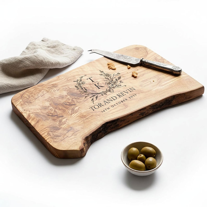 May include: A wooden serving board with a natural edge, engraved with floral details, initials, and the text "TOR AND KEVIN 16TH OCTOBER 2023." A cheese knife and a small bowl of olives are also present. A linen napkin is in the background.