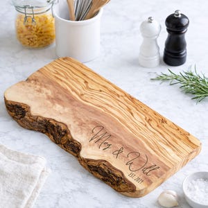 May include: A wooden cutting board with a natural edge. The board is engraved with the names "Meg & Will" and the date "EST. 2019". The board is light brown with darker wood grain patterns. The board is surrounded by kitchen items.