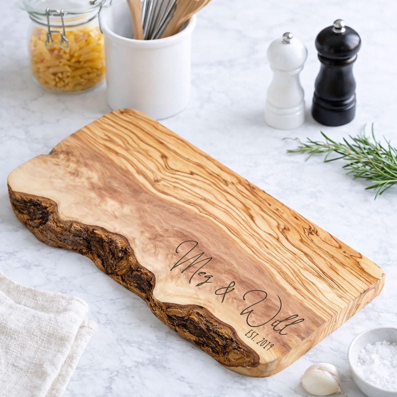 May include: A wooden cutting board with a natural edge. The board is engraved with the names "Meg & Will" and the date "EST. 2019". The board is light brown with darker wood grain patterns. The board is surrounded by kitchen items.