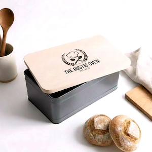 May include: A rectangular, gray bread box with a light wood lid. The lid features the text "THE RUSTIC OVEN EST. 2023" with a chef's hat and crossed utensils logo. Two round loaves of bread sit in front of the box.