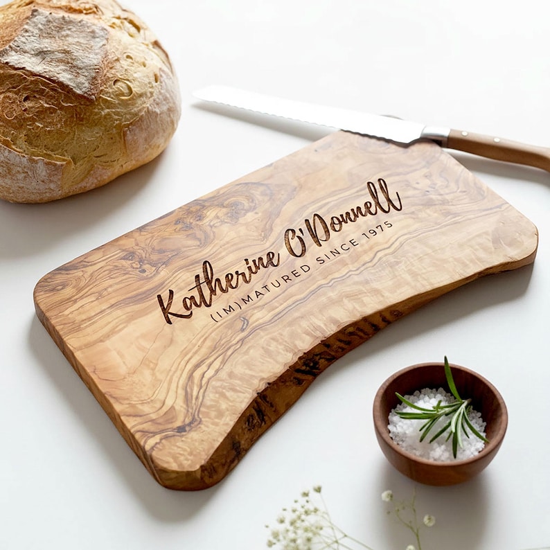 May include: A wooden cutting board with the engraved text "Katherine O'Donnell (I'M) MATURED SINCE 1975". A loaf of bread, a bread knife, and a small wooden bowl with salt and rosemary are also present.