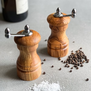 May include: Two wooden salt and pepper grinders with stainless steel handles. The grinders are made of light brown wood with a natural grain pattern. Black peppercorns and white salt are scattered around the grinders.