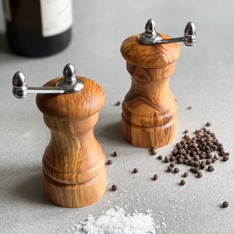 May include: Two wooden salt and pepper grinders with stainless steel handles. The grinders are made of light brown wood with a natural grain pattern. Black peppercorns and white salt are scattered around the grinders.