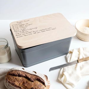 以下が含まれることがあります： 明るい木製の蓋が付いた灰色のパン箱。蓋には手書きのかぼちゃパンのレシピがあります。パンのパン、パンナイフ、液体の瓶が箱の隣にあります。小麦粉のボウルも画像にあります。