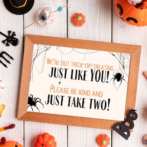 May include: A printable sign with a brown frame and white background. The sign says "We're out trick-or-treating just like you! Please be kind and just take two!" with a spider web design and a black spider in the corner.