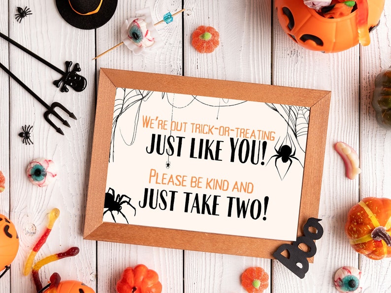 May include: A printable sign with a brown frame and white background. The sign says "We're out trick-or-treating just like you! Please be kind and just take two!" with a spider web design and a black spider in the corner.