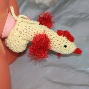 May include: A crocheted rooster toy with red and yellow accents. The rooster is wearing a yellow crocheted hat and has a red pom-pom on top.
