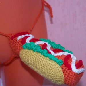 May include: A crocheted hot dog costume with a yellow bun, red ketchup, white mayonnaise, and green lettuce.
