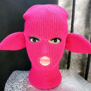May include: A bright hot pink knit balaclava featuring two prominent, pointed ear-like extensions on the sides. The headgear is constructed with a ribbed texture and includes three distinct openings for the eyes and the mouth area.