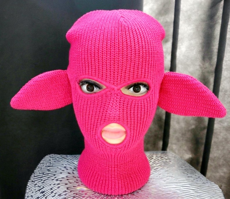 May include: A bright hot pink knit balaclava featuring two prominent, pointed ear-like extensions on the sides. The headgear is constructed with a ribbed texture and includes three distinct openings for the eyes and the mouth area.