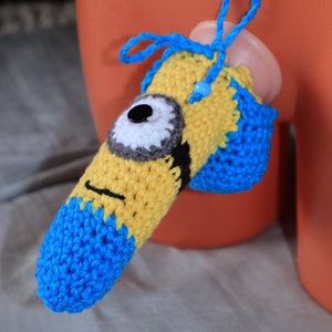 Op de afbeelding: Een geel en blauw gehaakte fallische hoes met één wit oog met een zwarte knoop als pupil, een zwarte geborduurde mond en een blauw trekkoord met een kleine blauwe kraal aan de bovenkant, ontworpen om over een cilindervormig object te passen.