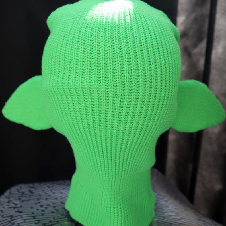 May include: A bright neon green knit balaclava featuring a ribbed texture throughout the entire piece. The head covering includes two prominent, pointed ear-like extensions protruding from the sides, crafted from the same vibrant green yarn.