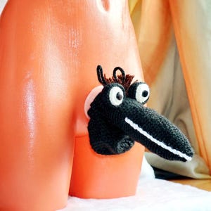 May include: A black and white crocheted creature with large eyes and a long snout is attached to an orange surface. The creature has brown hair-like details and is positioned to the side. The background is a soft, neutral color.