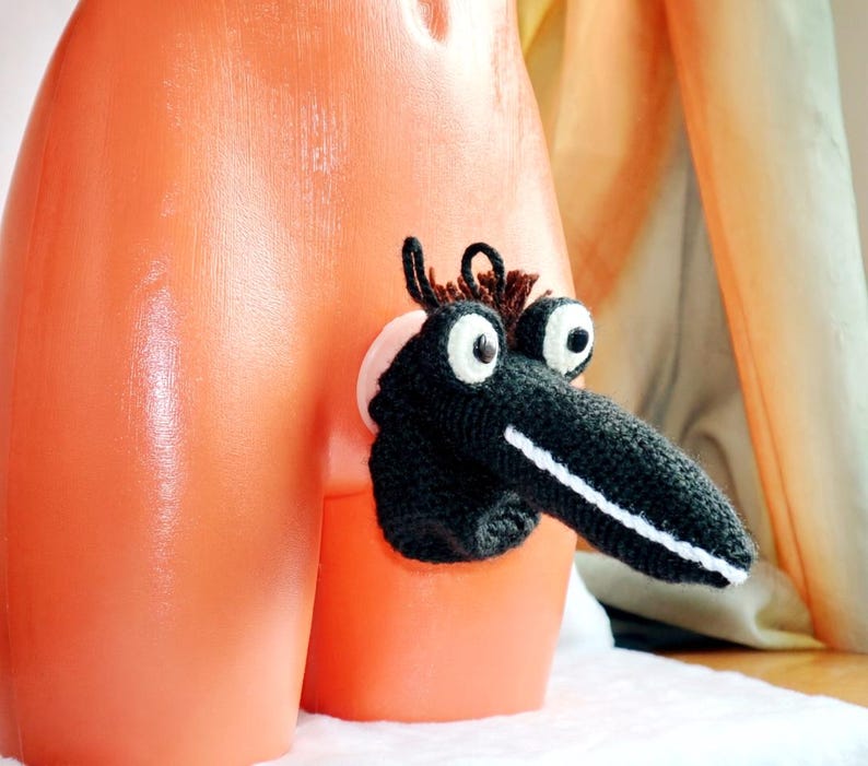 May include: A black and white crocheted creature with large eyes and a long snout is attached to an orange surface. The creature has brown hair-like details and is positioned to the side. The background is a soft, neutral color.