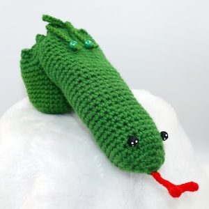 May include: Green crocheted snake with black eyes and a red tongue. The snake is lying on a white fluffy surface.