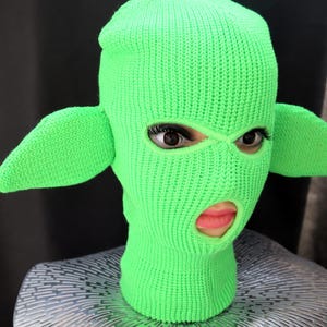 May include: A bright neon green knit balaclava featuring two large, pointed, ear-like protrusions extending from the sides. The head covering has two eye openings and a single mouth opening, revealing a face underneath the knitted fabric.