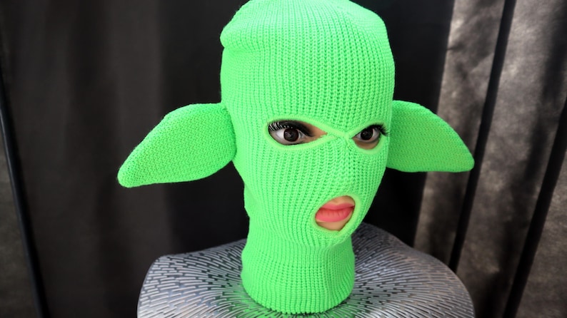 May include: A bright neon green knit balaclava featuring two large, pointed, ear-like protrusions extending from the sides. The head covering has two eye openings and a single mouth opening, revealing a face underneath the knitted fabric.