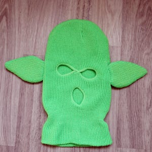 May include: A bright neon green knit balaclava featuring two pointed, ear-like extensions on the sides. The headgear has a ribbed bottom edge and three distinct openings: two for the eyes and one for the mouth, all finished with matching green trim.