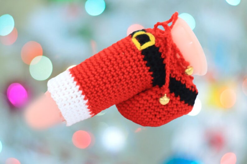 May include: A red and white crocheted Santa Claus costume for a small object. The costume has a black belt with a yellow buckle and gold jingle bells.