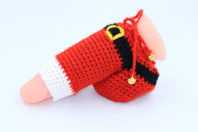 May include: A crocheted Santa Claus costume for a small object, featuring a red and white hat, a red suit with a black belt, and gold jingle bells.