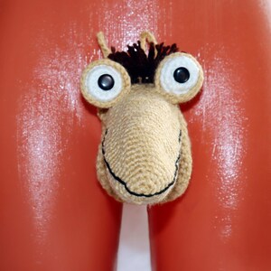 May include: A novelty crochet item in the shape of a cartoonish face with large, round eyes and a smiling mouth. The item is beige with black and brown accents. The background is a glossy orange surface.