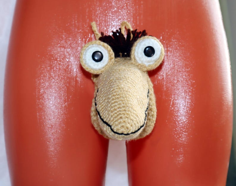 May include: A novelty crochet item in the shape of a cartoonish face with large, round eyes and a smiling mouth. The item is beige with black and brown accents. The background is a glossy orange surface.