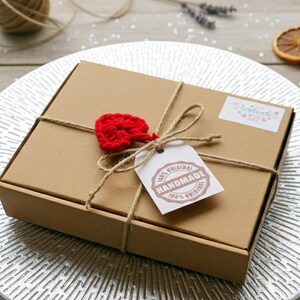 May include: A brown cardboard box tied with twine, adorned with a red crocheted heart and a tag that reads "100% Original Handmade." The box is on a silver plate, with dried orange slices and lavender sprigs in the background.
