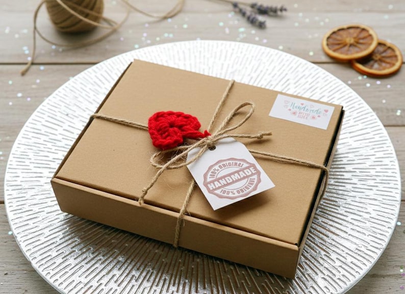 May include: A brown cardboard box tied with twine, adorned with a red crocheted heart and a tag that reads "100% Original Handmade." The box is on a silver plate, with dried orange slices and lavender sprigs in the background.
