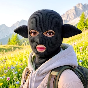 May include: A black knit balaclava featuring two pointed, ear-like protrusions on the sides. The headgear has a ribbed texture and includes openings for the eyes and mouth, covering the head and neck completely with a snug, fitted construction.