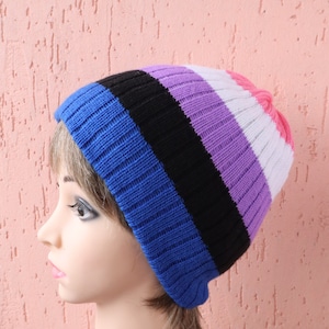 May include: A striped beanie hat in blue, black, purple, white, and pink. The hat is made of a ribbed knit material and is designed to be worn on the head. The colors are arranged in vertical stripes.