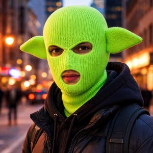 May include: A bright neon green knit balaclava featuring two prominent, pointed ears extending from the sides. The headgear has a ribbed texture and includes two circular openings for the eyes and a larger opening for the mouth area.