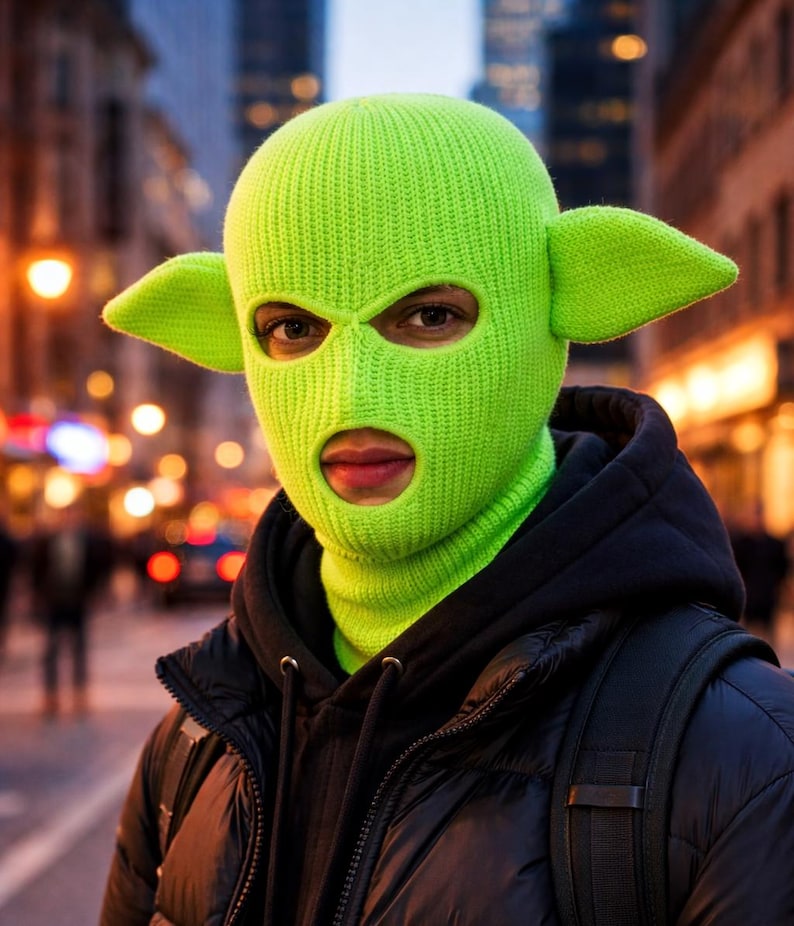 May include: A bright neon green knit balaclava featuring two prominent, pointed ears extending from the sides. The headgear has a ribbed texture and includes two circular openings for the eyes and a larger opening for the mouth area.