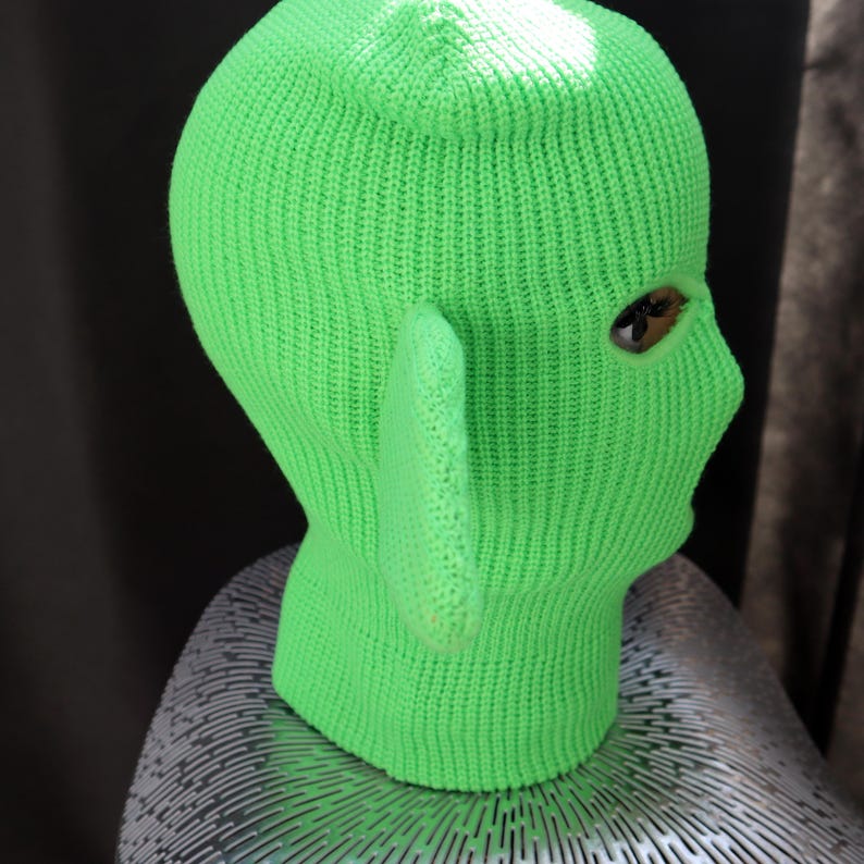 May include: A bright neon green knit balaclava featuring a ribbed texture and a single prominent, pointed ear-like extension on the side. The head covering includes a single visible eye opening and is constructed from a thick, uniform yarn material.