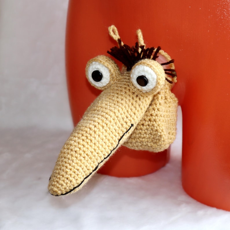 May include: A whimsical, handmade crochet creature with large, black-pupiled eyes, a long nose, and a tuft of brown hair. The creature is beige with black stitching details. It is attached to a red surface.