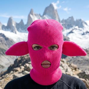 May include: A bright pink knit balaclava featuring two protruding, ear-like appendages on the sides. The headgear has a ribbed texture and includes two circular openings for the eyes and one larger circular opening for the mouth area.