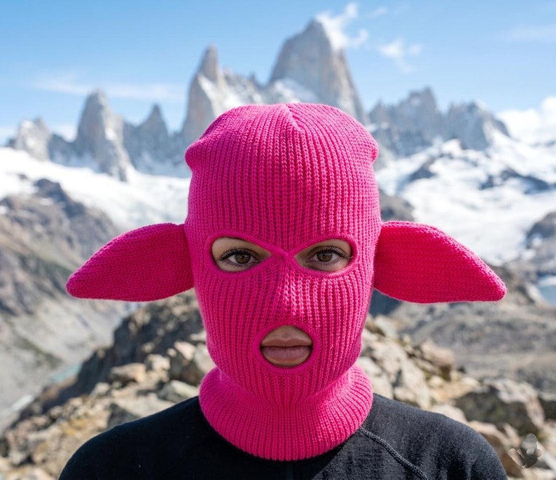May include: A bright pink knit balaclava featuring two protruding, ear-like appendages on the sides. The headgear has a ribbed texture and includes two circular openings for the eyes and one larger circular opening for the mouth area.
