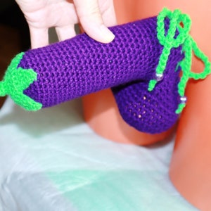 May include: A purple crocheted aubergine with a green stem and a green crocheted loop. The aubergine is being held by a hand.