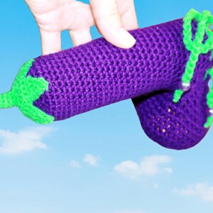 May include: A crocheted aubergine toy with green accents and beads. The aubergine is purple and has a textured surface.