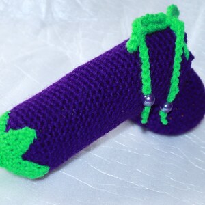 May include: A crocheted aubergine toy with green leaves and a purple body. The aubergine has two green stems with silver beads.