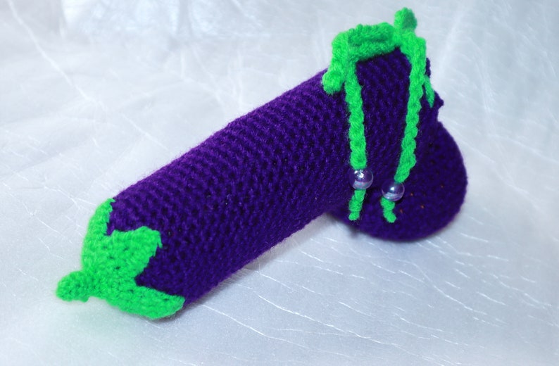 May include: A crocheted aubergine toy with green leaves and a purple body. The aubergine has two green stems with silver beads.