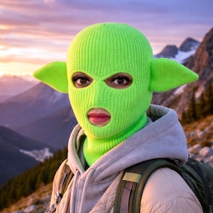 May include: A bright neon green knit balaclava featuring two prominent, pointed ears extending from the sides. The headgear has two eye openings and a single mouth opening, crafted from a ribbed knit fabric that covers the head and neck area.