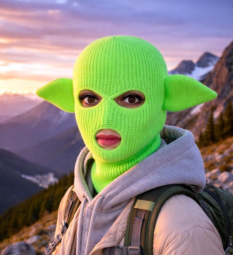 May include: A bright neon green knit balaclava featuring two prominent, pointed ears extending from the sides. The headgear has two eye openings and a single mouth opening, crafted from a ribbed knit fabric that covers the head and neck area.