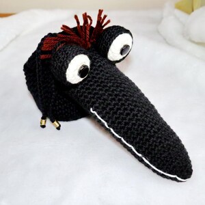 May include: A handmade black crochet creature with large white-rimmed eyes and a long beak. The creature has brown yarn hair and gold-colored accents. A white line outlines the beak.