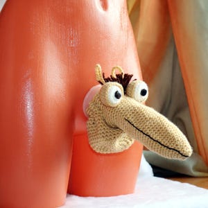 May include: A whimsical, handmade crochet creature with large, googly eyes and a long, pointed nose. The creature is tan and brown, attached to an orange plastic form. The design is playful and unique, showcasing a creative craft project.