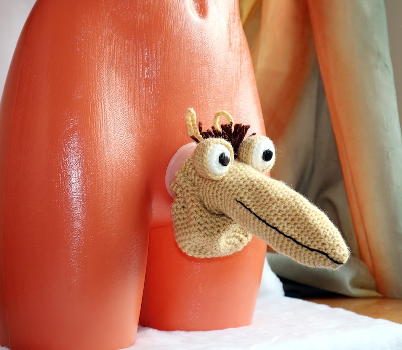 May include: A whimsical, handmade crochet creature with large, googly eyes and a long, pointed nose. The creature is tan and brown, attached to an orange plastic form. The design is playful and unique, showcasing a creative craft project.