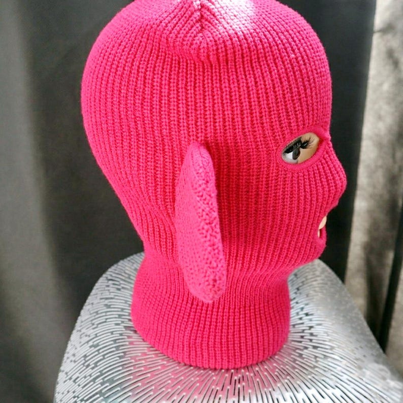 May include: A bright hot pink knit balaclava featuring a ribbed texture and a single prominent ear-like protrusion on the side. The head covering includes a single oval-shaped eye opening and a separate mouth opening, crafted from a thick yarn material.