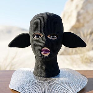 May include: A black knitted balaclava featuring two prominent, pointed ear-like extensions on the sides. The headgear covers the entire head and neck, with cutouts for the eyes and mouth, revealing a face with dark purple lipstick underneath.
