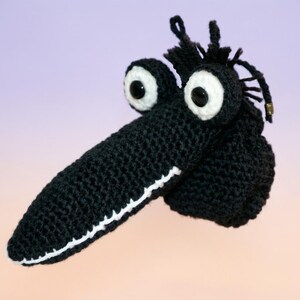 May include: A black crocheted novelty item with large white-rimmed eyes and a long, pointed snout. The item has a white stitched border along the snout and a tuft of black yarn on top. The background is a gradient of purple and peach.