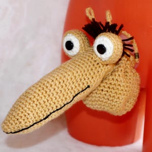 May include: A whimsical, handmade crochet creature with a long, tan snout, large white-rimmed eyes with black pupils, and a small tuft of dark brown hair. The creature is attached to an orange surface.